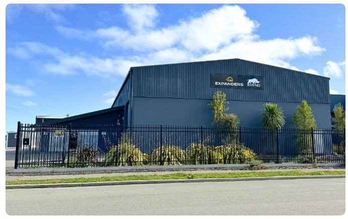 Industrial building with blue exterior and logos "Expanders" and "Rhyno" on facade, surrounded by a black metal fence and plants, under a blue sky with clouds.